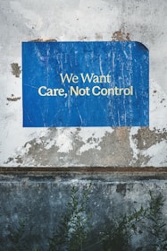 A weathered and textured wall with a vivid blue poster attached. The poster bears the words 'We Want Care, Not Control' in bold white capital letters. Visible below the poster are patches of chipped paint and rough concrete. Dark green foliage grows at the base, adding a natural element to the scene.