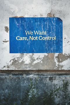 A weathered and textured wall with a vivid blue poster attached. The poster bears the words 'We Want Care, Not Control' in bold white capital letters. Visible below the poster are patches of chipped paint and rough concrete. Dark green foliage grows at the base, adding a natural element to the scene.