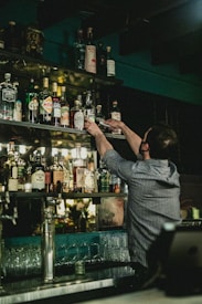 A bartender is reaching up to grab a bottle from a well-stocked shelf full of various spirits and liquors. The setting appears to be a dimly lit bar with bottles of different shapes and sizes arranged neatly. Glasses are stacked below the shelf, and a cash register is visible in the lower right corner.