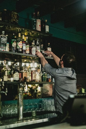 A bartender is reaching up to grab a bottle from a well-stocked shelf full of various spirits and liquors. The setting appears to be a dimly lit bar with bottles of different shapes and sizes arranged neatly. Glasses are stacked below the shelf, and a cash register is visible in the lower right corner.