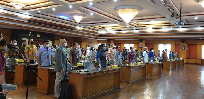 A formal gathering in a large, well-lit conference room with ornate chandeliers on the ceiling. Rows of desks are occupied by individuals standing in formal attire, many wearing face masks. There is a microphone stand in the foreground and electronic devices on the tables.