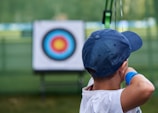 A young person wearing a blue cap aims a bow and arrow towards a target in the distance. The target has concentric circles in blue, red, and yellow colors, and is placed outdoors in a green, grassy area.