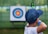 A young person wearing a blue cap aims a bow and arrow towards a target in the distance. The target has concentric circles in blue, red, and yellow colors, and is placed outdoors in a green, grassy area.