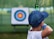 A young person wearing a blue cap aims a bow and arrow towards a target in the distance. The target has concentric circles in blue, red, and yellow colors, and is placed outdoors in a green, grassy area.