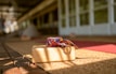 A wooden yoga block with a colorful, patterned yoga strap is placed on a woven mat. Sunlight casts shadows across the scene, creating a warm and inviting atmosphere. The background shows blurred yoga mats and a spacious area with wooden flooring, suggesting a yoga or meditation space.