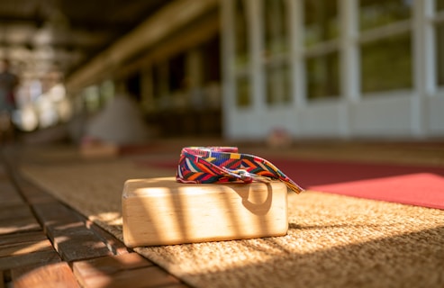 A wooden yoga block with a colorful, patterned yoga strap is placed on a woven mat. Sunlight casts shadows across the scene, creating a warm and inviting atmosphere. The background shows blurred yoga mats and a spacious area with wooden flooring, suggesting a yoga or meditation space.
