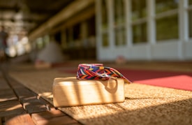 A wooden yoga block with a colorful, patterned yoga strap is placed on a woven mat. Sunlight casts shadows across the scene, creating a warm and inviting atmosphere. The background shows blurred yoga mats and a spacious area with wooden flooring, suggesting a yoga or meditation space.