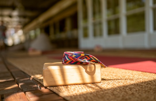 A wooden yoga block with a colorful, patterned yoga strap is placed on a woven mat. Sunlight casts shadows across the scene, creating a warm and inviting atmosphere. The background shows blurred yoga mats and a spacious area with wooden flooring, suggesting a yoga or meditation space.