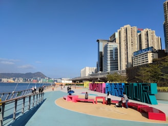 A vibrant cityscape with tall modern buildings under a clear blue sky. A waterfront promenade features colorful, large sculptural letters spelling out a word, with people walking and interacting in this public space. The calm water and distant mountains add depth to the background.