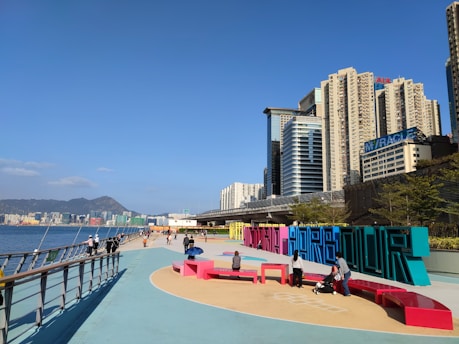 A vibrant cityscape with tall modern buildings under a clear blue sky. A waterfront promenade features colorful, large sculptural letters spelling out a word, with people walking and interacting in this public space. The calm water and distant mountains add depth to the background.