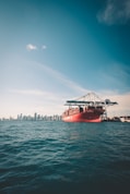 A large red cargo ship carrying shipping containers is situated in the foreground on the ocean. In the background, a city skyline can be seen with tall buildings under a clear blue sky.