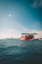 A large red cargo ship carrying shipping containers is situated in the foreground on the ocean. In the background, a city skyline can be seen with tall buildings under a clear blue sky.