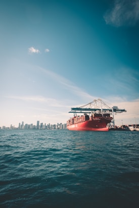 A large red cargo ship carrying shipping containers is situated in the foreground on the ocean. In the background, a city skyline can be seen with tall buildings under a clear blue sky.