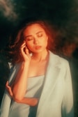 A woman with closed eyes is bathed in warm, golden light, creating a serene and dreamy atmosphere. Her hand gently touches her face, and she is dressed in a light-colored blazer over a matching top.