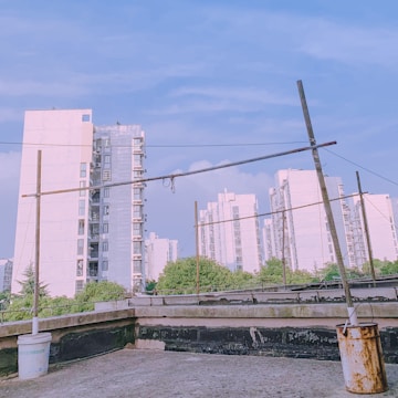 Several high-rise residential buildings are in the background, with a clear blue sky above. The foreground features a rooftop area with makeshift poles and wires, along with a white bucket and a rusted can.