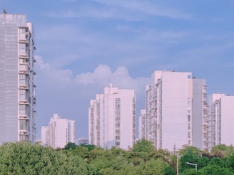 Several modern high-rise apartment buildings are surrounded by lush green trees under a clear blue sky. The buildings are mostly white, contrasting with the greenery below, and there are a few clouds visible in the sky.