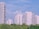 Several modern high-rise apartment buildings are surrounded by lush green trees under a clear blue sky. The buildings are mostly white, contrasting with the greenery below, and there are a few clouds visible in the sky.