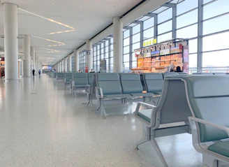 An empty airport terminal with rows of empty seating and large windows. There is a food or retail kiosk visible in the background, with bright signage and products displayed. The flooring appears to be a smooth, light-colored surface and the environment is well-lit with natural light coming from outside.