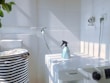 A white laundry area with a washing machine featuring a Samsung logo. On top of the machine is a blue spray bottle. To the left, there is a striped laundry basket. The walls are tiled, and there is a hose attached to the wall. Sunlight casts shadows creating a grid pattern on the surfaces.