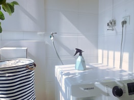 A white laundry area with a washing machine featuring a Samsung logo. On top of the machine is a blue spray bottle. To the left, there is a striped laundry basket. The walls are tiled, and there is a hose attached to the wall. Sunlight casts shadows creating a grid pattern on the surfaces.