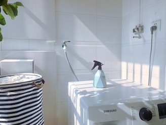A white laundry area with a washing machine featuring a Samsung logo. On top of the machine is a blue spray bottle. To the left, there is a striped laundry basket. The walls are tiled, and there is a hose attached to the wall. Sunlight casts shadows creating a grid pattern on the surfaces.