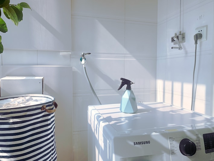 A white laundry area with a washing machine featuring a Samsung logo. On top of the machine is a blue spray bottle. To the left, there is a striped laundry basket. The walls are tiled, and there is a hose attached to the wall. Sunlight casts shadows creating a grid pattern on the surfaces.