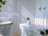 A white laundry area with a washing machine featuring a Samsung logo. On top of the machine is a blue spray bottle. To the left, there is a striped laundry basket. The walls are tiled, and there is a hose attached to the wall. Sunlight casts shadows creating a grid pattern on the surfaces.