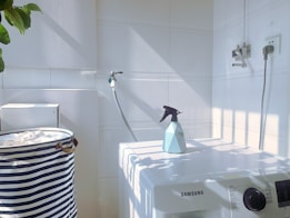 A white laundry area with a washing machine featuring a Samsung logo. On top of the machine is a blue spray bottle. To the left, there is a striped laundry basket. The walls are tiled, and there is a hose attached to the wall. Sunlight casts shadows creating a grid pattern on the surfaces.