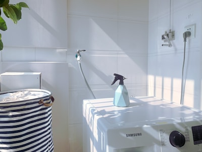 A white laundry area with a washing machine featuring a Samsung logo. On top of the machine is a blue spray bottle. To the left, there is a striped laundry basket. The walls are tiled, and there is a hose attached to the wall. Sunlight casts shadows creating a grid pattern on the surfaces.
