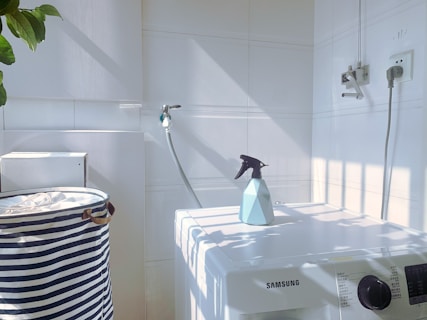A white laundry area with a washing machine featuring a Samsung logo. On top of the machine is a blue spray bottle. To the left, there is a striped laundry basket. The walls are tiled, and there is a hose attached to the wall. Sunlight casts shadows creating a grid pattern on the surfaces.