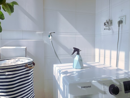 A white laundry area with a washing machine featuring a Samsung logo. On top of the machine is a blue spray bottle. To the left, there is a striped laundry basket. The walls are tiled, and there is a hose attached to the wall. Sunlight casts shadows creating a grid pattern on the surfaces.