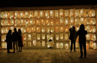 A group of people stand in front of a wall adorned with many illuminated paper lanterns. The lanterns are arranged in neat rows and emit a warm, soft glow, creating a serene and enchanting atmosphere. The silhouettes of a few individuals suggest they are observing the lanterns closely, as if engaged in conversation or contemplation.
