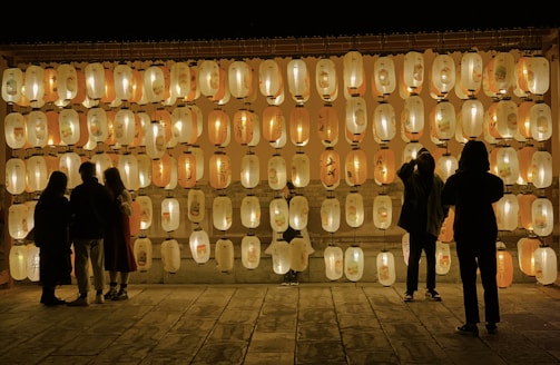 A group of people stand in front of a wall adorned with many illuminated paper lanterns. The lanterns are arranged in neat rows and emit a warm, soft glow, creating a serene and enchanting atmosphere. The silhouettes of a few individuals suggest they are observing the lanterns closely, as if engaged in conversation or contemplation.
