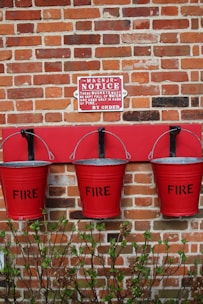 Three red fire buckets hanging on a brick wall beneath a notice sign. The buckets are labeled with the word 'FIRE' and are outlined against a strip of red paint on the wall. There is a small hedge with green leaves at the base of the wall.