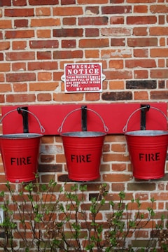 Three red fire buckets hanging on a brick wall beneath a notice sign. The buckets are labeled with the word 'FIRE' and are outlined against a strip of red paint on the wall. There is a small hedge with green leaves at the base of the wall.