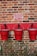 Three red fire buckets hanging on a brick wall beneath a notice sign. The buckets are labeled with the word 'FIRE' and are outlined against a strip of red paint on the wall. There is a small hedge with green leaves at the base of the wall.