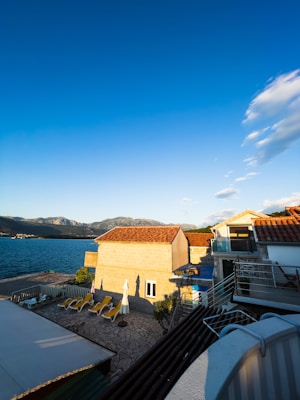 A seaside scene with a small courtyard area featuring several sun loungers and an umbrella. The foreground includes rooftops with red tiles and a stone building casting shadows. Beyond the building, the sea is visible against a backdrop of distant mountains under a clear blue sky with a few clouds.