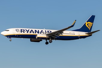 A commercial airplane with the Ryanair logo and branding is flying mid-air against a clear blue sky. It is a Boeing 737, predominantly white with blue and yellow accents.