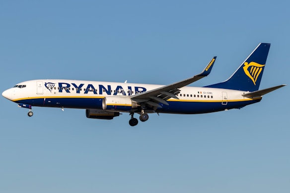 A commercial airplane with the Ryanair logo and branding is flying mid-air against a clear blue sky. It is a Boeing 737, predominantly white with blue and yellow accents.
