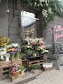 A charming flower shop displays various types of plants and flowers arranged on wooden crates and shelves outside a storefront. The facade features rustic decorations with leafy vines and a pink neon sign. On the pavement, a small model of the Eiffel Tower adds a unique touch, alongside chalkboard signs with floral prices. The area is adorned with a variety of colorful blooms, including roses, daisies, and potted plants.