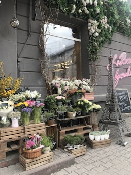 A charming flower shop displays various types of plants and flowers arranged on wooden crates and shelves outside a storefront. The facade features rustic decorations with leafy vines and a pink neon sign. On the pavement, a small model of the Eiffel Tower adds a unique touch, alongside chalkboard signs with floral prices. The area is adorned with a variety of colorful blooms, including roses, daisies, and potted plants.