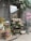 A charming flower shop displays various types of plants and flowers arranged on wooden crates and shelves outside a storefront. The facade features rustic decorations with leafy vines and a pink neon sign. On the pavement, a small model of the Eiffel Tower adds a unique touch, alongside chalkboard signs with floral prices. The area is adorned with a variety of colorful blooms, including roses, daisies, and potted plants.