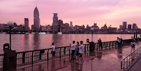 A vibrant urban skyline at dusk with skyscrapers reflecting on the calm river. The sky exhibits a mixture of pink and orange hues, contributing to a serene atmosphere. Several people are present on the riverside promenade, some standing and others engaged in conversation or taking photos.