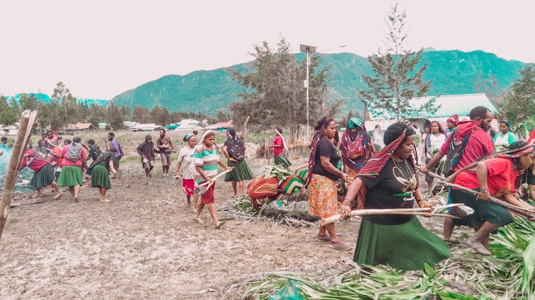 A group of people are participating in a communal activity outdoors, possibly a cultural or traditional event. They are dressed in colorful clothing, with several wearing skirts, headscarves, and jewelry. The setting is a natural landscape with mountains and trees in the background, indicating a rural or village environment.