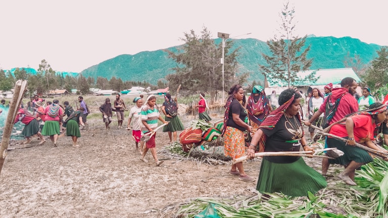 A group of people are participating in a communal activity outdoors, possibly a cultural or traditional event. They are dressed in colorful clothing, with several wearing skirts, headscarves, and jewelry. The setting is a natural landscape with mountains and trees in the background, indicating a rural or village environment.