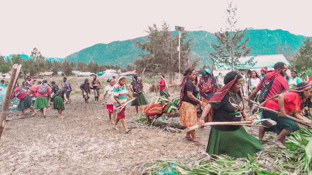 A group of people are participating in a communal activity outdoors, possibly a cultural or traditional event. They are dressed in colorful clothing, with several wearing skirts, headscarves, and jewelry. The setting is a natural landscape with mountains and trees in the background, indicating a rural or village environment.