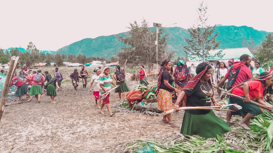 A group of people are participating in a communal activity outdoors, possibly a cultural or traditional event. They are dressed in colorful clothing, with several wearing skirts, headscarves, and jewelry. The setting is a natural landscape with mountains and trees in the background, indicating a rural or village environment.