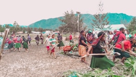 A group of people are participating in a communal activity outdoors, possibly a cultural or traditional event. They are dressed in colorful clothing, with several wearing skirts, headscarves, and jewelry. The setting is a natural landscape with mountains and trees in the background, indicating a rural or village environment.