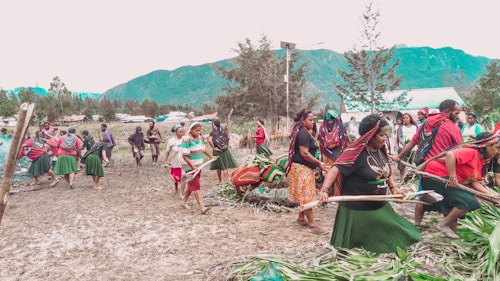 A group of people are participating in a communal activity outdoors, possibly a cultural or traditional event. They are dressed in colorful clothing, with several wearing skirts, headscarves, and jewelry. The setting is a natural landscape with mountains and trees in the background, indicating a rural or village environment.