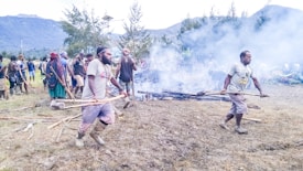 A group of people are engaged in an outdoor fire-walking ritual. Two men in the foreground walk across smoldering embers carrying long wooden poles. Smoke billows around them as others, dressed in traditional attire, observe or participate in the background. The scene is set in a natural environment with mountains and trees.